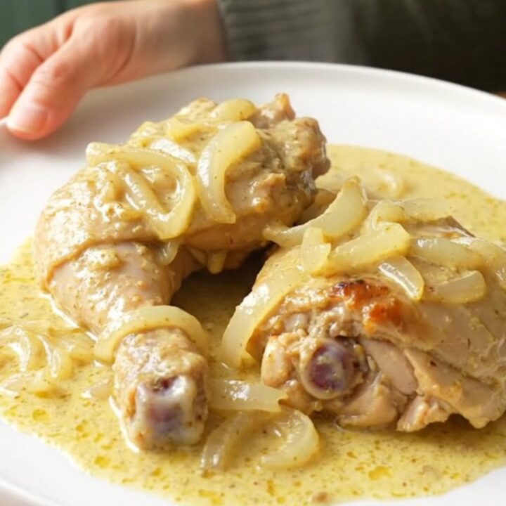 A stainless steel slow cooker filled with tender chicken thighs, soft onions, and a rich, creamy sauce.