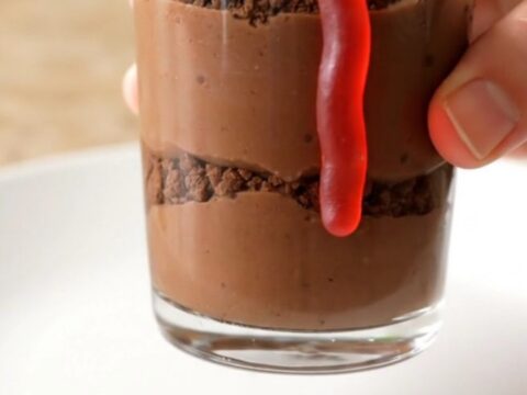 A dozen clear glass cups filled with chocolate pudding, topped with dark cookie crumbs and gummy worms on a baking sheet.
