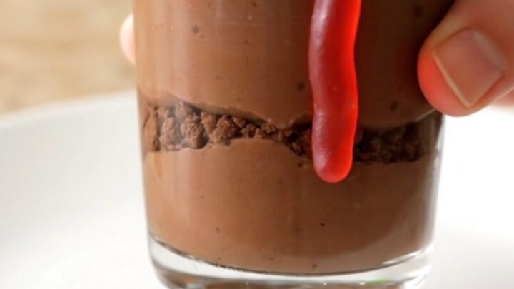 A dozen clear glass cups filled with chocolate pudding, topped with dark cookie crumbs and gummy worms on a baking sheet.