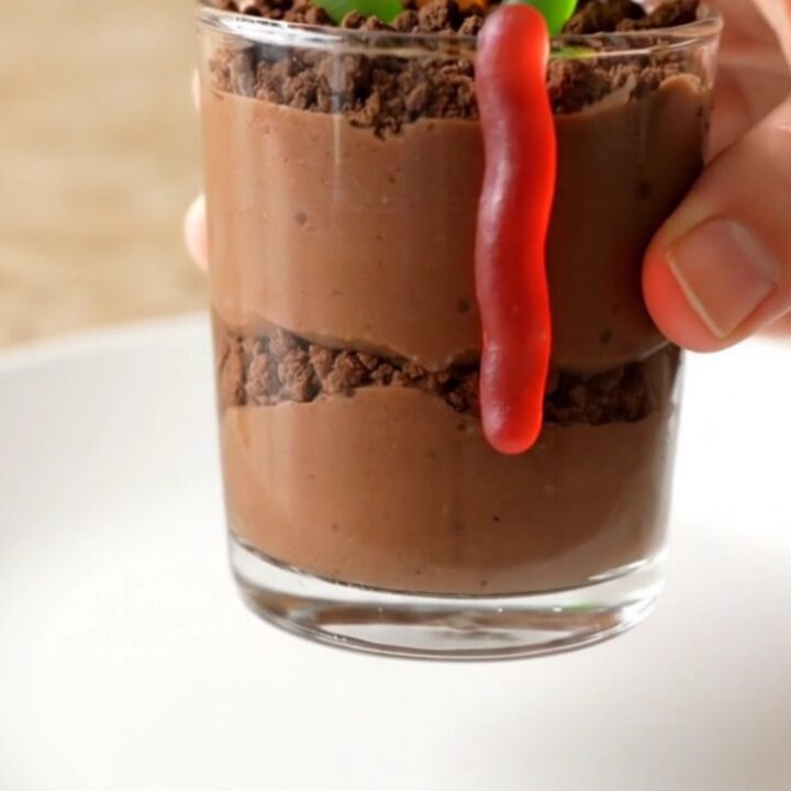 A dozen clear glass cups filled with chocolate pudding, topped with dark cookie crumbs and gummy worms on a baking sheet.