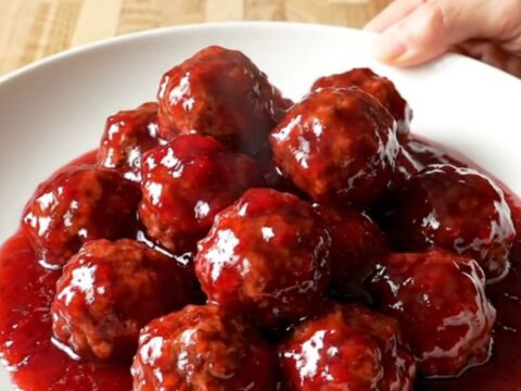 A stainless steel slow cooker filled with meatballs covered in a thick, glossy red raspberry glaze.