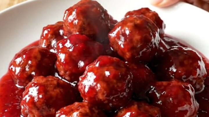A stainless steel slow cooker filled with meatballs covered in a thick, glossy red raspberry glaze.