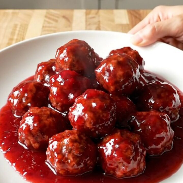 A stainless steel slow cooker filled with meatballs covered in a thick, glossy red raspberry glaze.