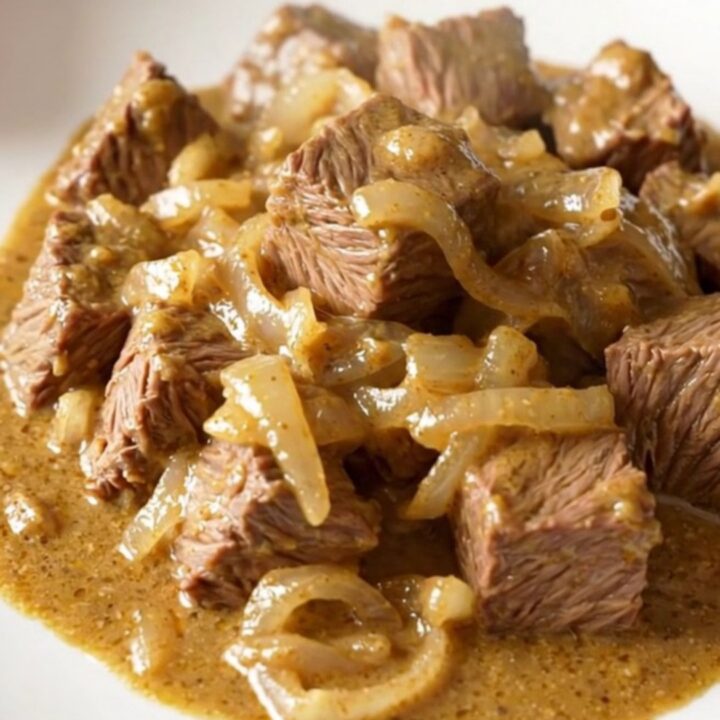 A close-up of tender beef cubes covered in a rich, creamy onion sauce served on a white plate.