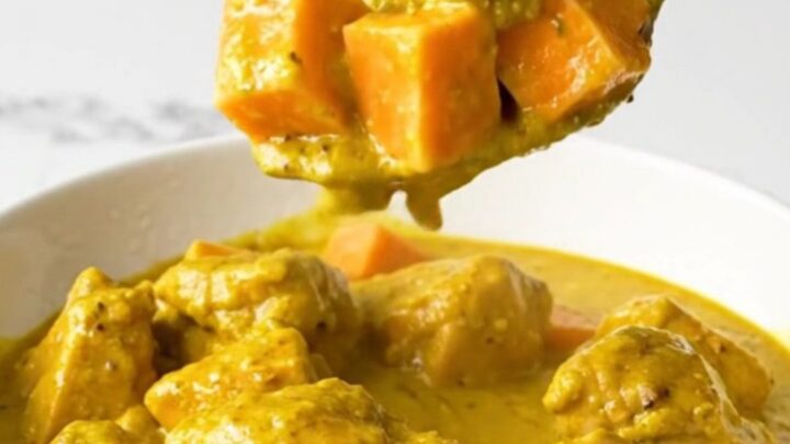 A top-down view of a black slow cooker filled with chicken and sweet potatoes in a golden coconut curry sauce.