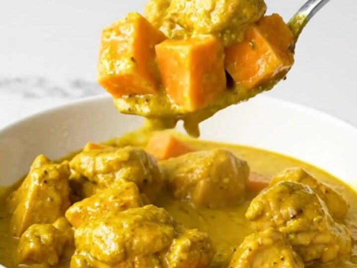 A top-down view of a black slow cooker filled with chicken and sweet potatoes in a golden coconut curry sauce.