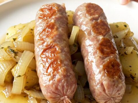 A stainless steel slow cooker filled with bratwurst, halved baby potatoes, and onions in a rich herb butter sauce.