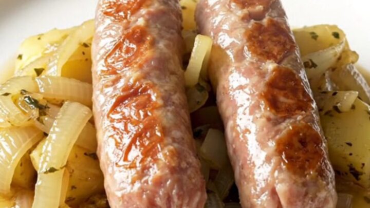 A stainless steel slow cooker filled with bratwurst, halved baby potatoes, and onions in a rich herb butter sauce.