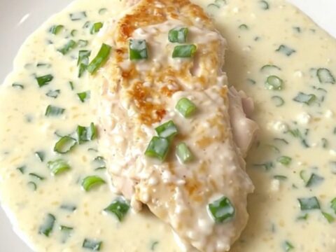 A close-up of a tender chicken breast covered in a thick, creamy white sauce with green onions, served on a white plate.