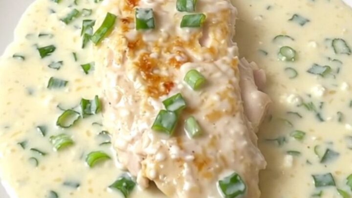 A close-up of a tender chicken breast covered in a thick, creamy white sauce with green onions, served on a white plate.