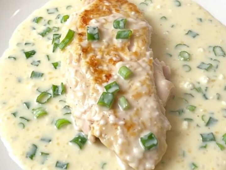 A close-up of a tender chicken breast covered in a thick, creamy white sauce with green onions, served on a white plate.