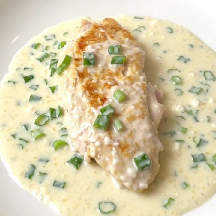 A close-up of a tender chicken breast covered in a thick, creamy white sauce with green onions, served on a white plate.