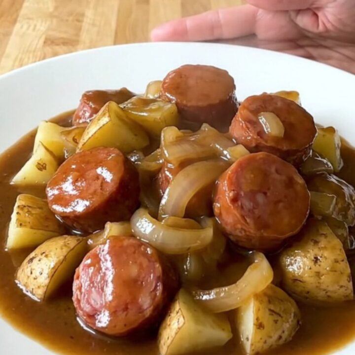 A close-up of a slow cooker filled with sliced smoked sausage and tender potato cubes in a rich brown gravy.