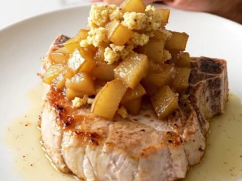 A tender, braised boneless pork chop topped with spiced apples and savory stuffing on a white plate.