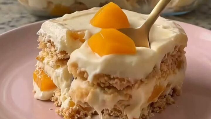 A close-up of a gold spoon scooping a layered peach and cream dessert from a pastel pink plate.