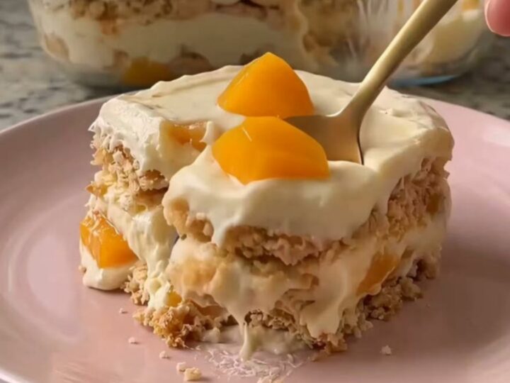 A close-up of a gold spoon scooping a layered peach and cream dessert from a pastel pink plate.