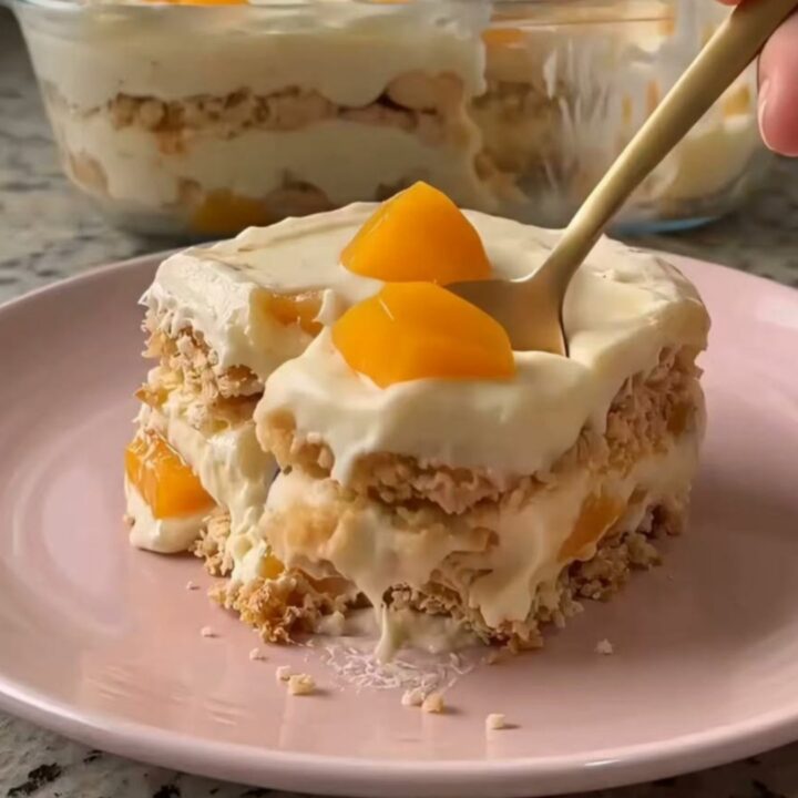 A close-up of a gold spoon scooping a layered peach and cream dessert from a pastel pink plate.