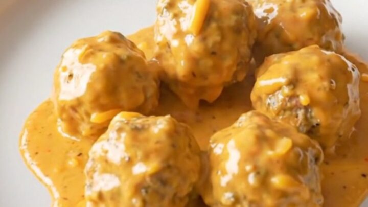 A stainless steel slow cooker filled with creamy meatballs in a rich, orange-tinted cheese sauce.