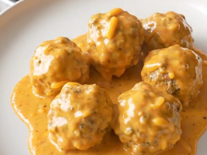A stainless steel slow cooker filled with creamy meatballs in a rich, orange-tinted cheese sauce.