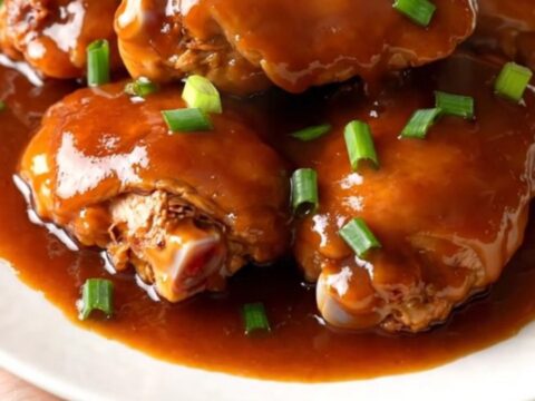 A top-down view of cooked chicken thighs in a slow cooker coated in a glossy red-brown sauce with green onions.