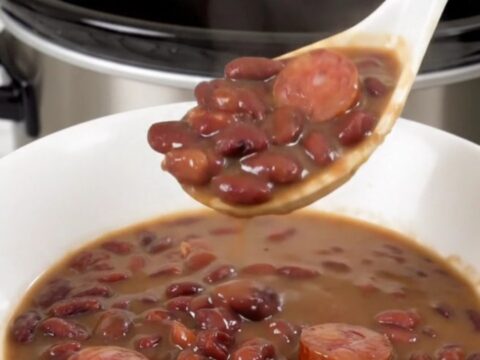A top-down view of a black slow cooker filled with red beans and sliced smoked sausage.