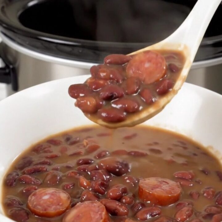 A top-down view of a black slow cooker filled with red beans and sliced smoked sausage.