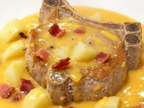 A plate featuring a tender pork chop covered in a creamy cheddar cheese sauce with potatoes and bacon.