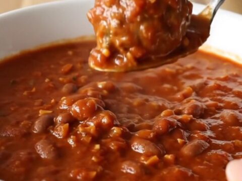A white bowl filled with thick, hearty meatball chili, garnished with a spoon, set against a blurred kitchen background.