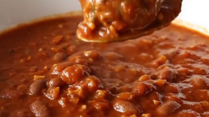 A white bowl filled with thick, hearty meatball chili, garnished with a spoon, set against a blurred kitchen background.