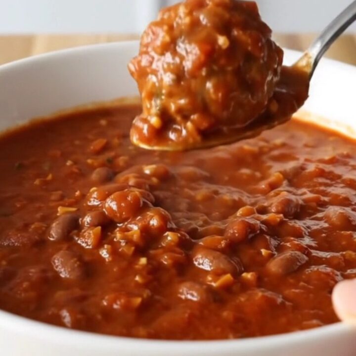 A white bowl filled with thick, hearty meatball chili, garnished with a spoon, set against a blurred kitchen background.