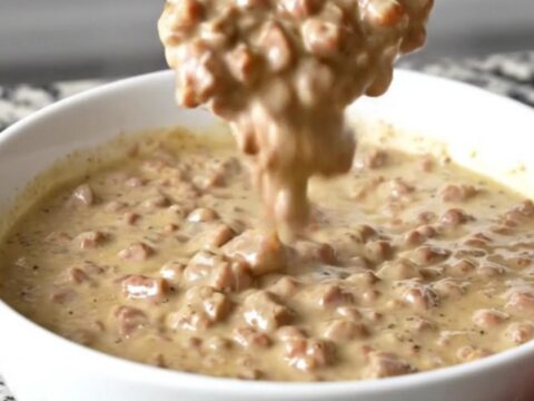 A close-up of a creamy, peppery sausage gravy dip in a white bowl with a spoon, steam rising.