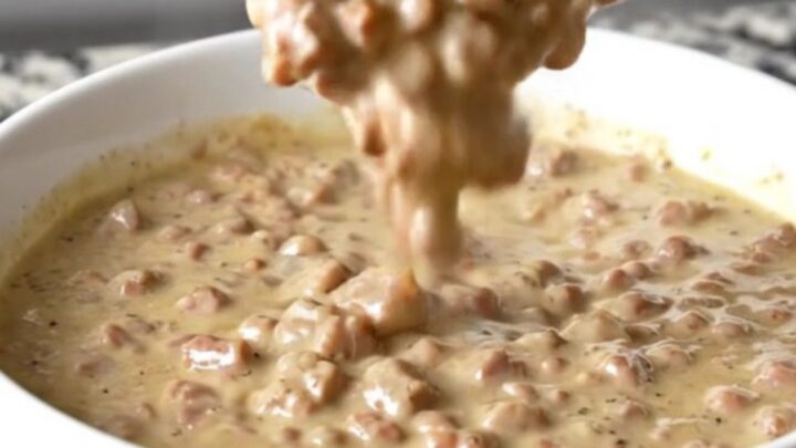 A close-up of a creamy, peppery sausage gravy dip in a white bowl with a spoon, steam rising.
