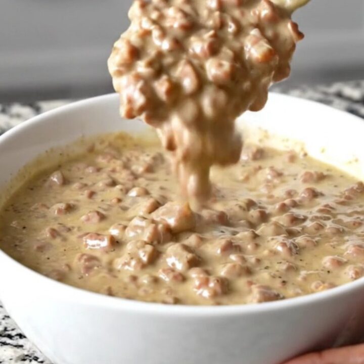 A close-up of a creamy, peppery sausage gravy dip in a white bowl with a spoon, steam rising.
