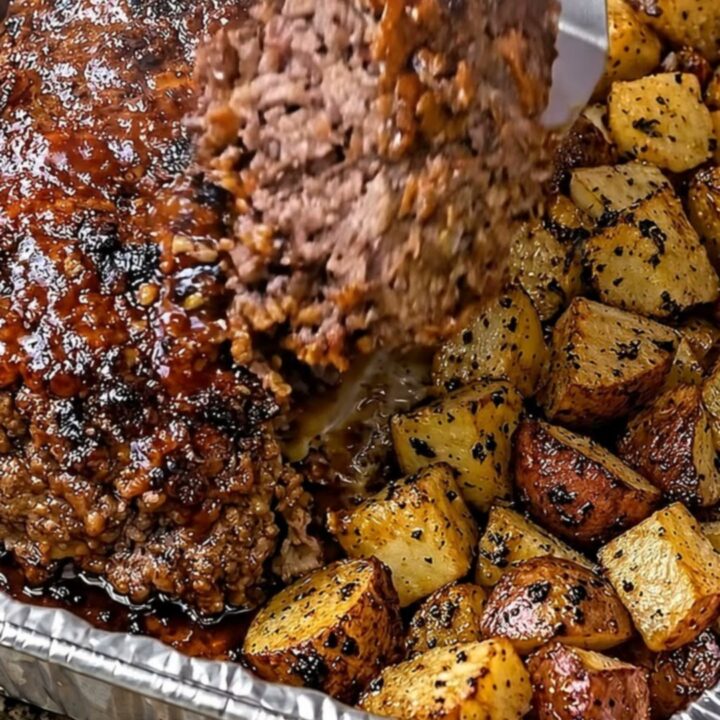 A steaming, juicy slice of glazed meatloaf served with crispy golden roasted potatoes in a foil pan.