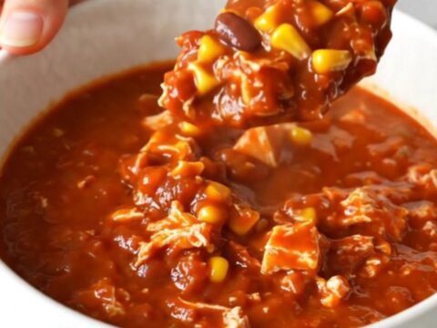 A top-down view of a black slow cooker filled with raw ground turkey, beans, corn, and red enchilada sauce.