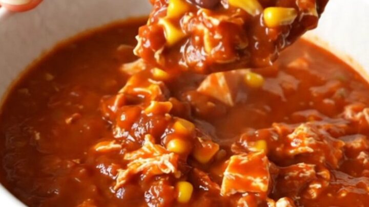A top-down view of a black slow cooker filled with raw ground turkey, beans, corn, and red enchilada sauce.