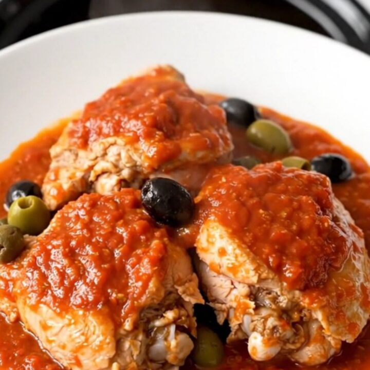 A top-down view of a black slow cooker filled with raw chicken thighs and a rich tomato sauce with olives.