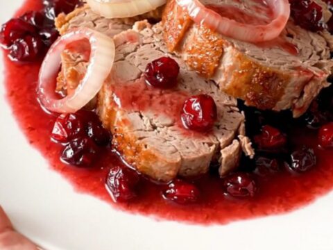 A slow cooker filled with a whole pork loin, onion rings, and a glossy cranberry sauce.