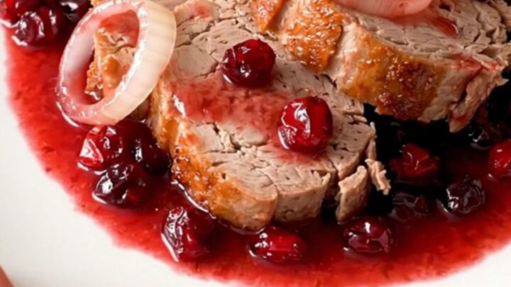 A slow cooker filled with a whole pork loin, onion rings, and a glossy cranberry sauce.