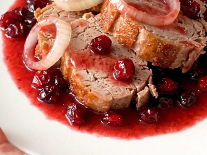 A slow cooker filled with a whole pork loin, onion rings, and a glossy cranberry sauce.