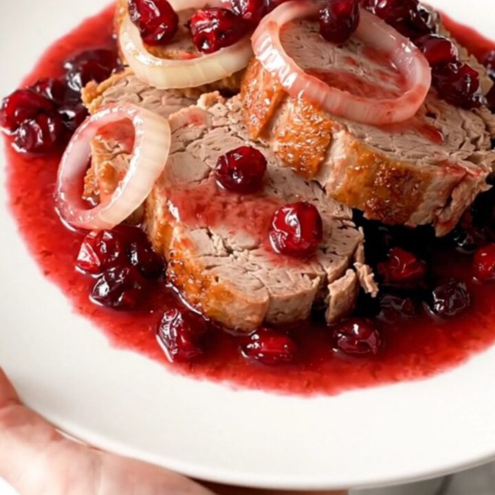 A slow cooker filled with a whole pork loin, onion rings, and a glossy cranberry sauce.