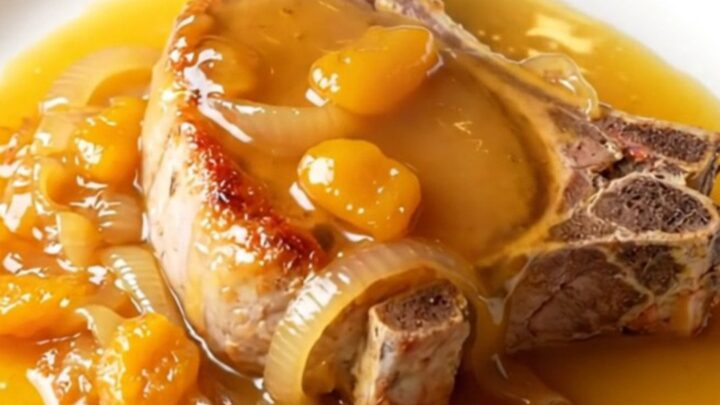 A close-up of a tender, slow-braised bone-in pork chop topped with soft onions and a glossy apricot glaze on a white plate.