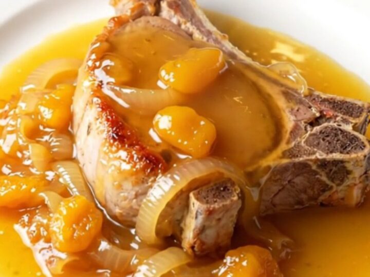 A close-up of a tender, slow-braised bone-in pork chop topped with soft onions and a glossy apricot glaze on a white plate.