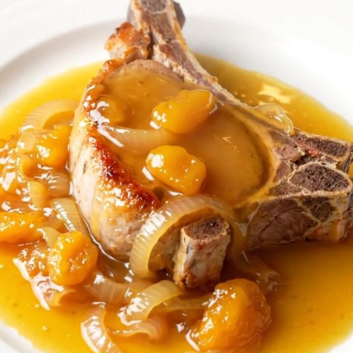 A close-up of a tender, slow-braised bone-in pork chop topped with soft onions and a glossy apricot glaze on a white plate.