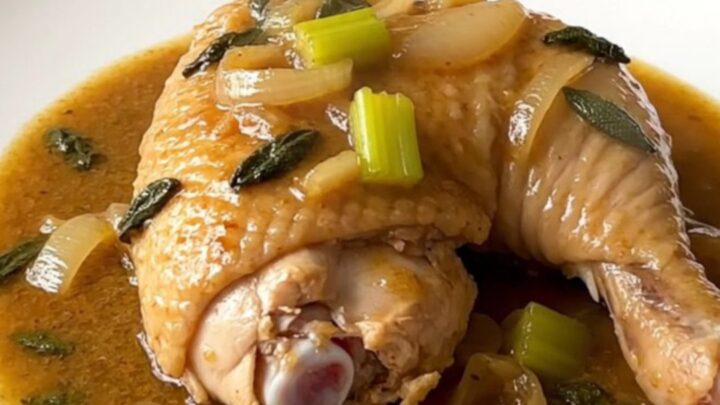 A close-up of tender, braised chicken leg quarters in a rich, sage-infused gravy in a white slow cooker.