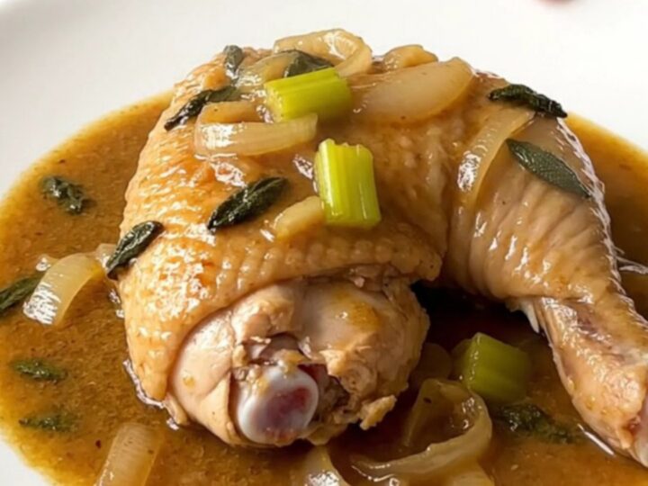 A close-up of tender, braised chicken leg quarters in a rich, sage-infused gravy in a white slow cooker.