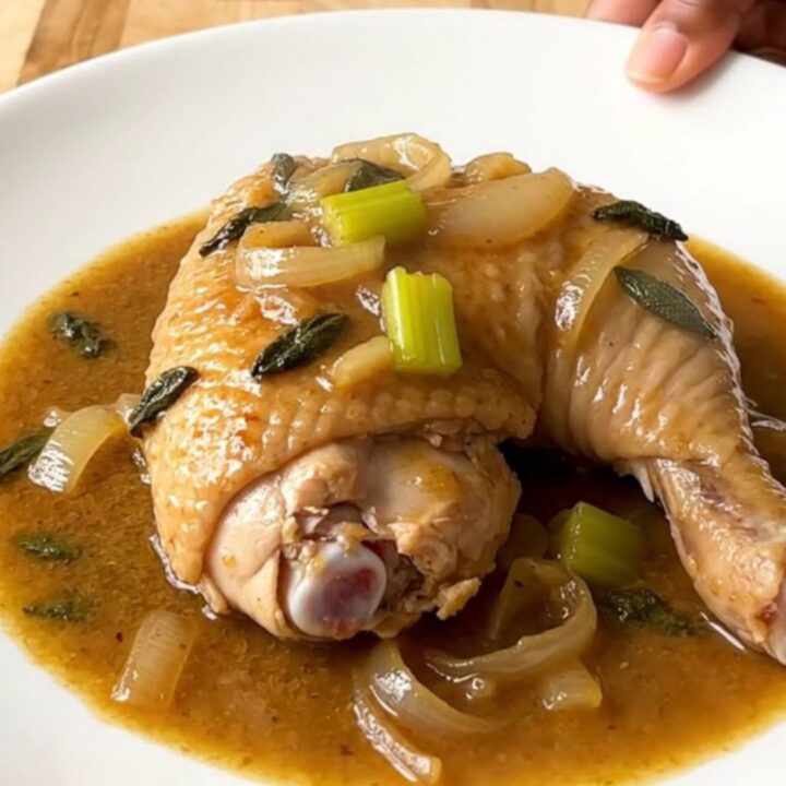 A close-up of tender, braised chicken leg quarters in a rich, sage-infused gravy in a white slow cooker.