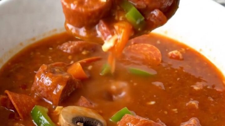 A top-down view of a black slow cooker filled with raw Italian sausage, pepperoni, peppers, and mushrooms.