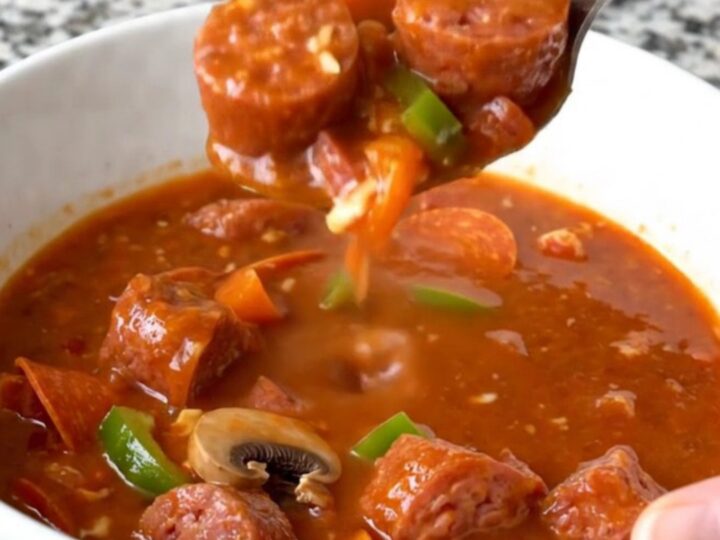 A top-down view of a black slow cooker filled with raw Italian sausage, pepperoni, peppers, and mushrooms.