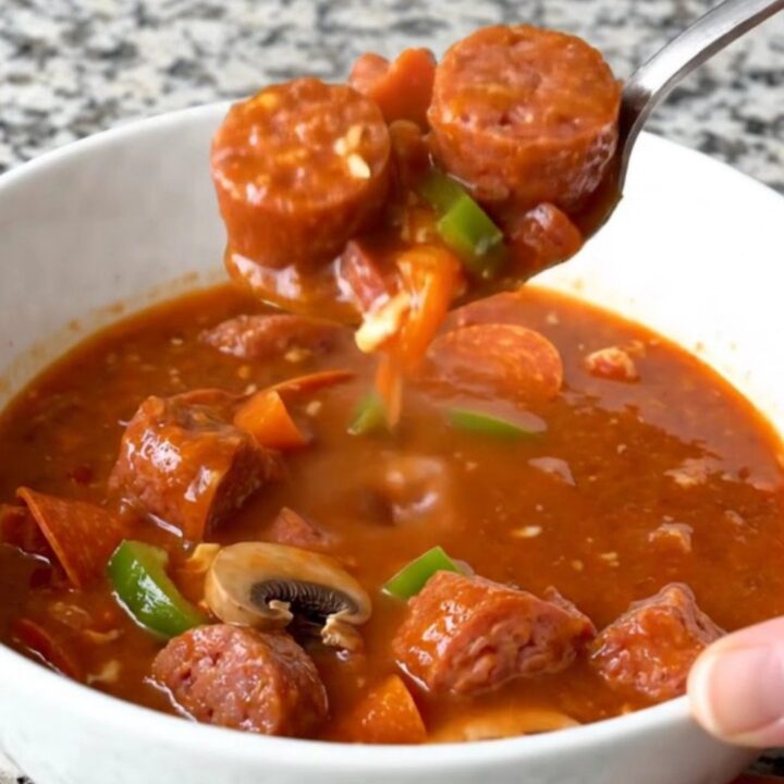 A top-down view of a black slow cooker filled with raw Italian sausage, pepperoni, peppers, and mushrooms.